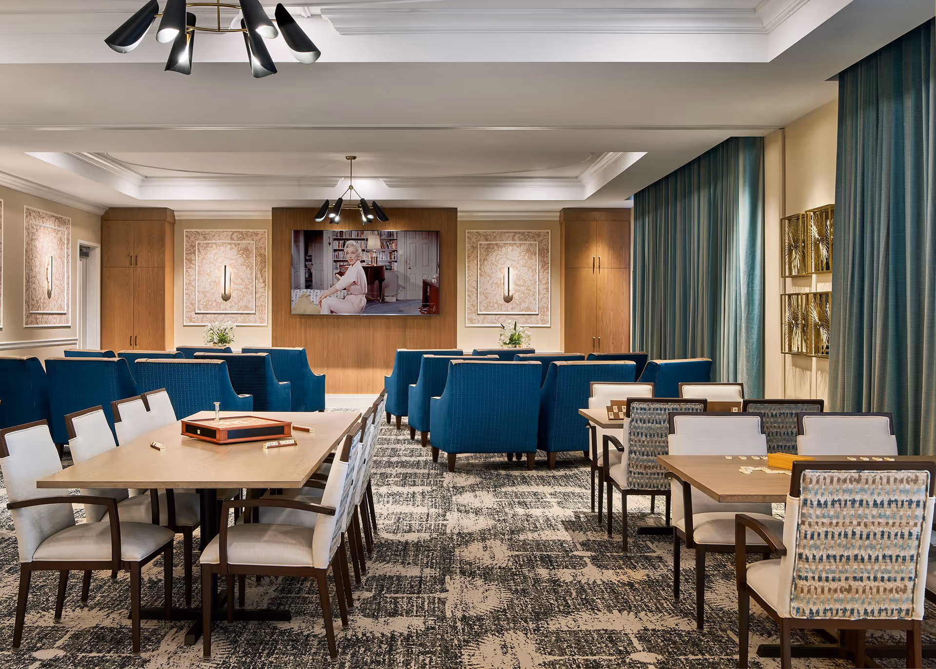 A well-lit common area with multiple tables and chairs arranged for group activities or dining. The room features blue upholstered armchairs facing a large wall-mounted television displaying a woman sitting in a room. The walls have decorative panels with sconces, and there are tall curtains on the right side. The carpet has a patterned design, and modern light fixtures hang from the ceiling.