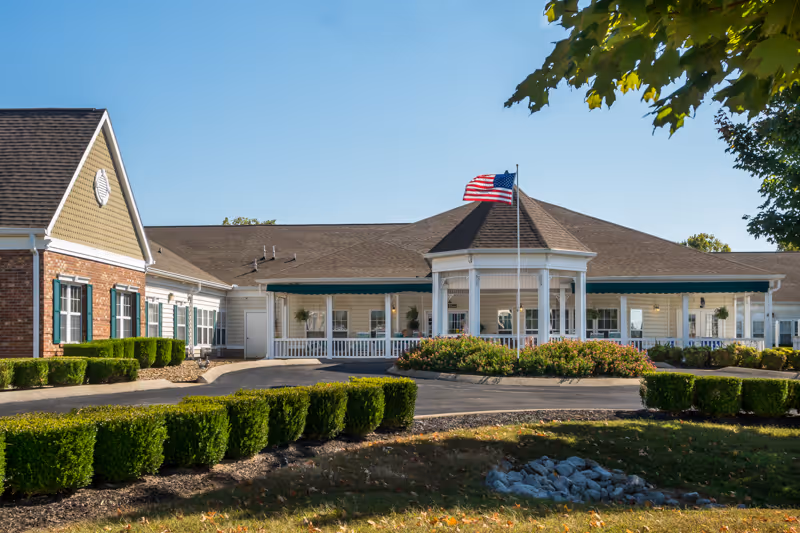 Front exterior view of a single-story senior living facility building with a peaked roof, white columns, and a circular driveway. An American flag is flying on a flagpole in front of the building, surrounded by neatly trimmed bushes and landscaping under a clear blue sky.