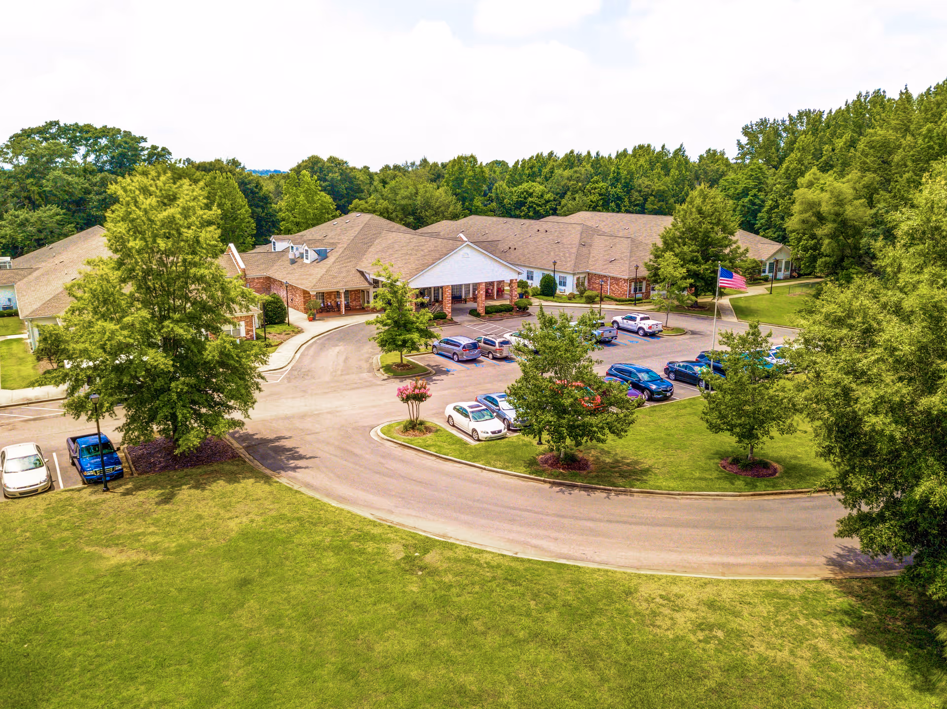 Aerial view of Bridgewood Gardens senior living facility showing a large single-story building with a brown roof surrounded by trees and greenery. There is a curved driveway with parked cars and an American flag near the entrance. The area is lush with grass and landscaped with small trees and bushes.