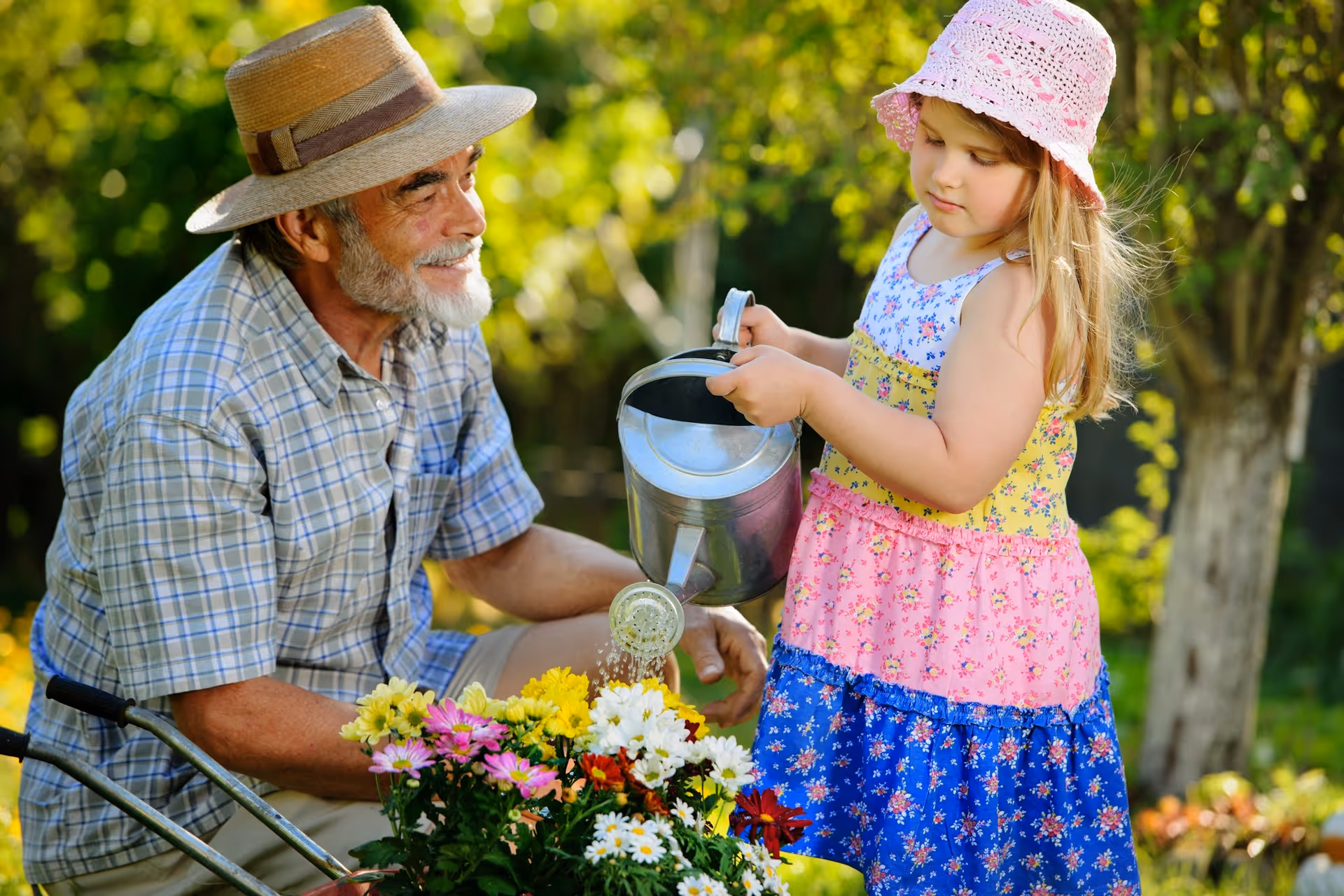 An elderly man wearing a straw hat and plaid shirt smiles while watching a young girl in a colorful dress and pink hat water flowers in a garden.