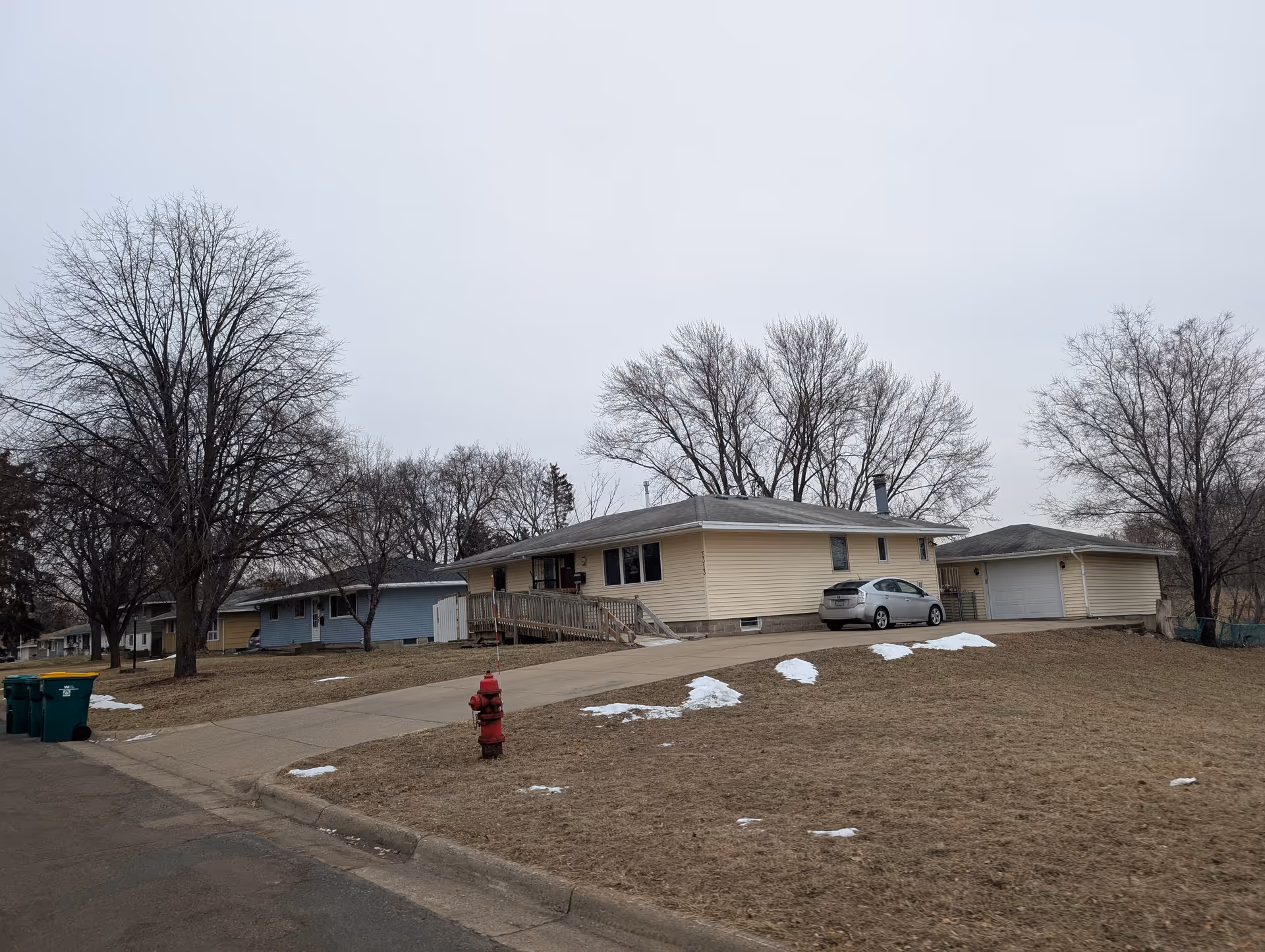 Single-story ranch-style house with a driveway, garage and parked car on a residential street with leafless trees and small patches of snow.