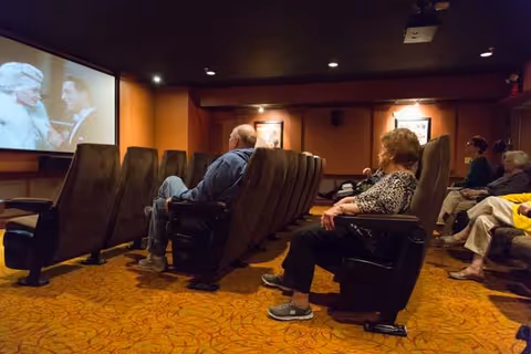 Several elderly people seated in comfortable theater-style chairs watching a movie projected on a screen in a dimly lit room with warm-colored walls and patterned carpet.