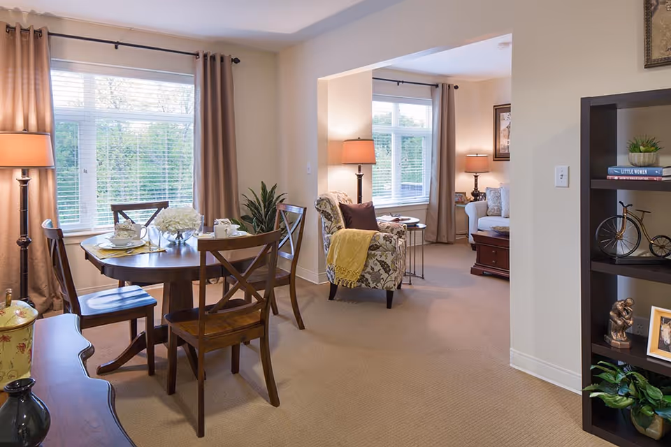 A cozy senior living interior featuring a dining area with a round wooden table set for two, surrounded by four wooden chairs. The room has large windows with beige curtains allowing natural light to fill the space. Adjacent to the dining area is a living room with a patterned armchair draped with a yellow throw blanket, a white sofa, side tables with lamps, and decorative items on a dark wooden bookshelf.