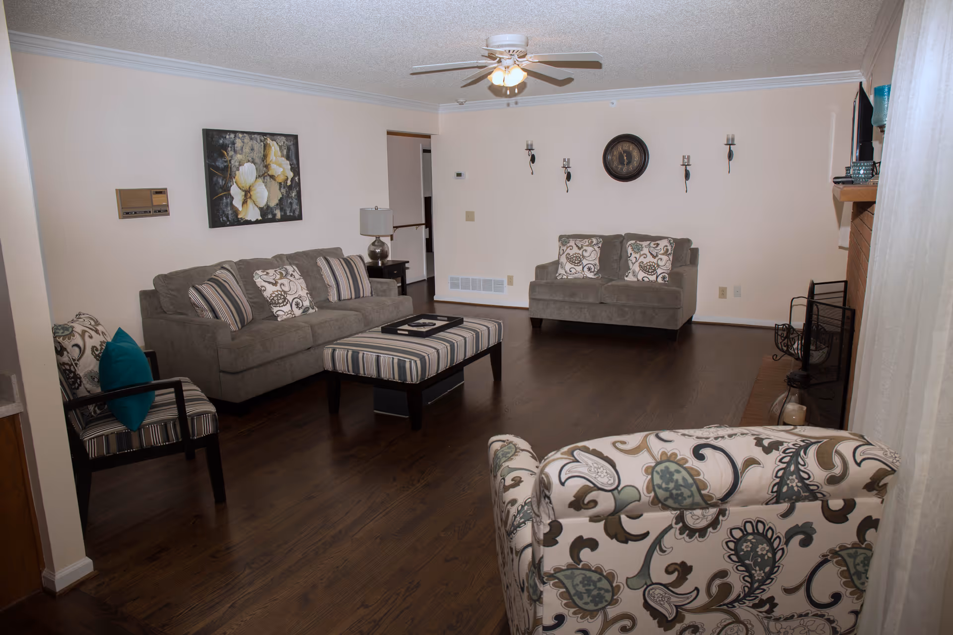 Spacious living room with gray sofas, a patterned armchair, striped ottoman, hardwood floor, wall decor, and a ceiling fan.