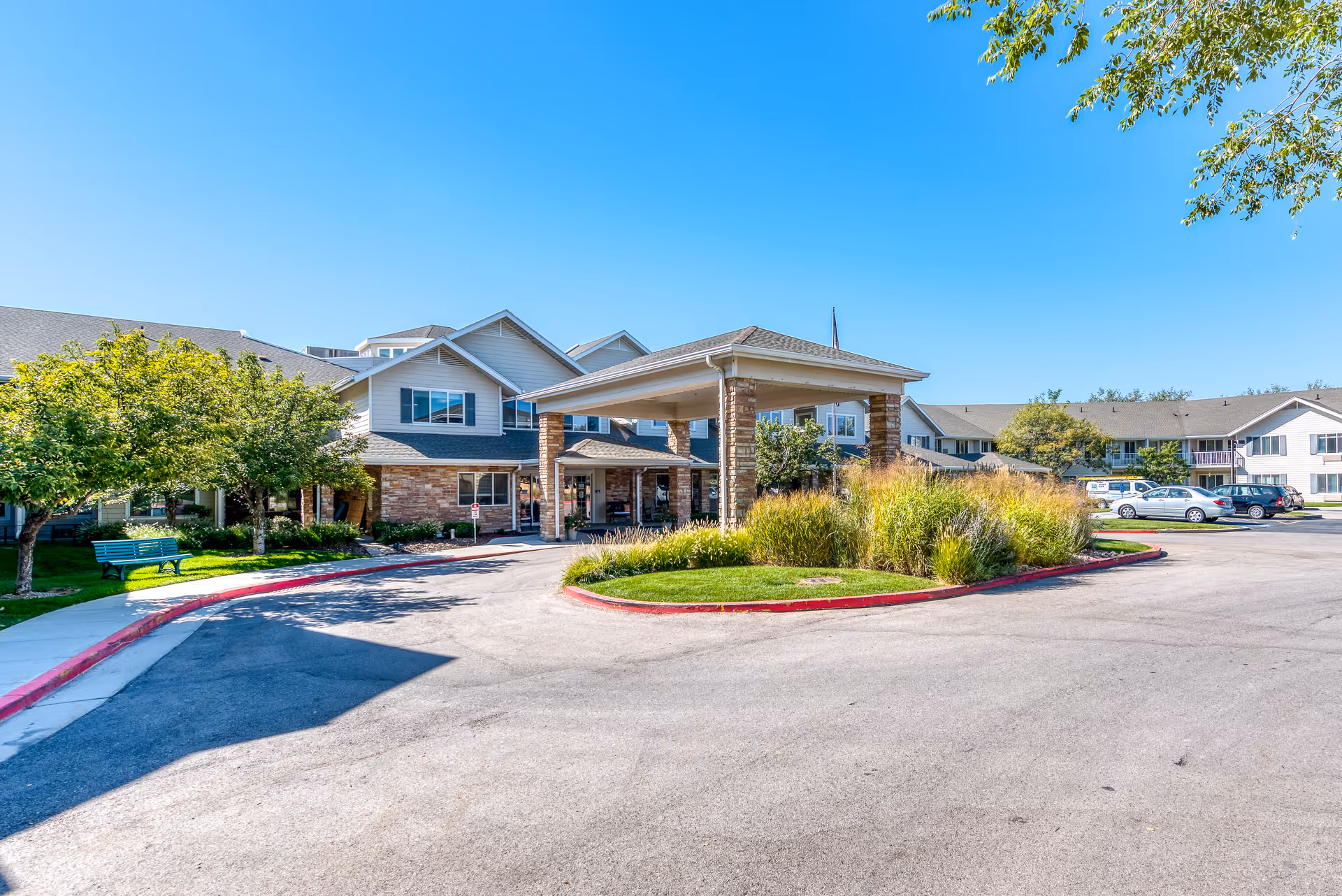 Exterior view of Solstice Senior Living at Sandy facility showing a large driveway with a covered entrance supported by stone pillars, surrounded by landscaped greenery and trees under a clear blue sky. Several parked cars are visible in the background.