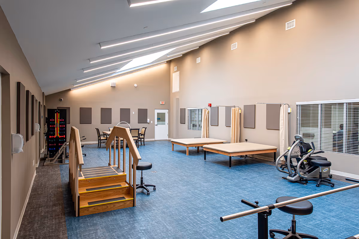 A rehabilitation therapy room with blue flooring and beige walls. The room features a wooden set of stairs with handrails, two therapy beds with privacy curtains, exercise equipment including a stationary bike, and a table with chairs in the background. The ceiling is slanted with skylights allowing natural light to enter.