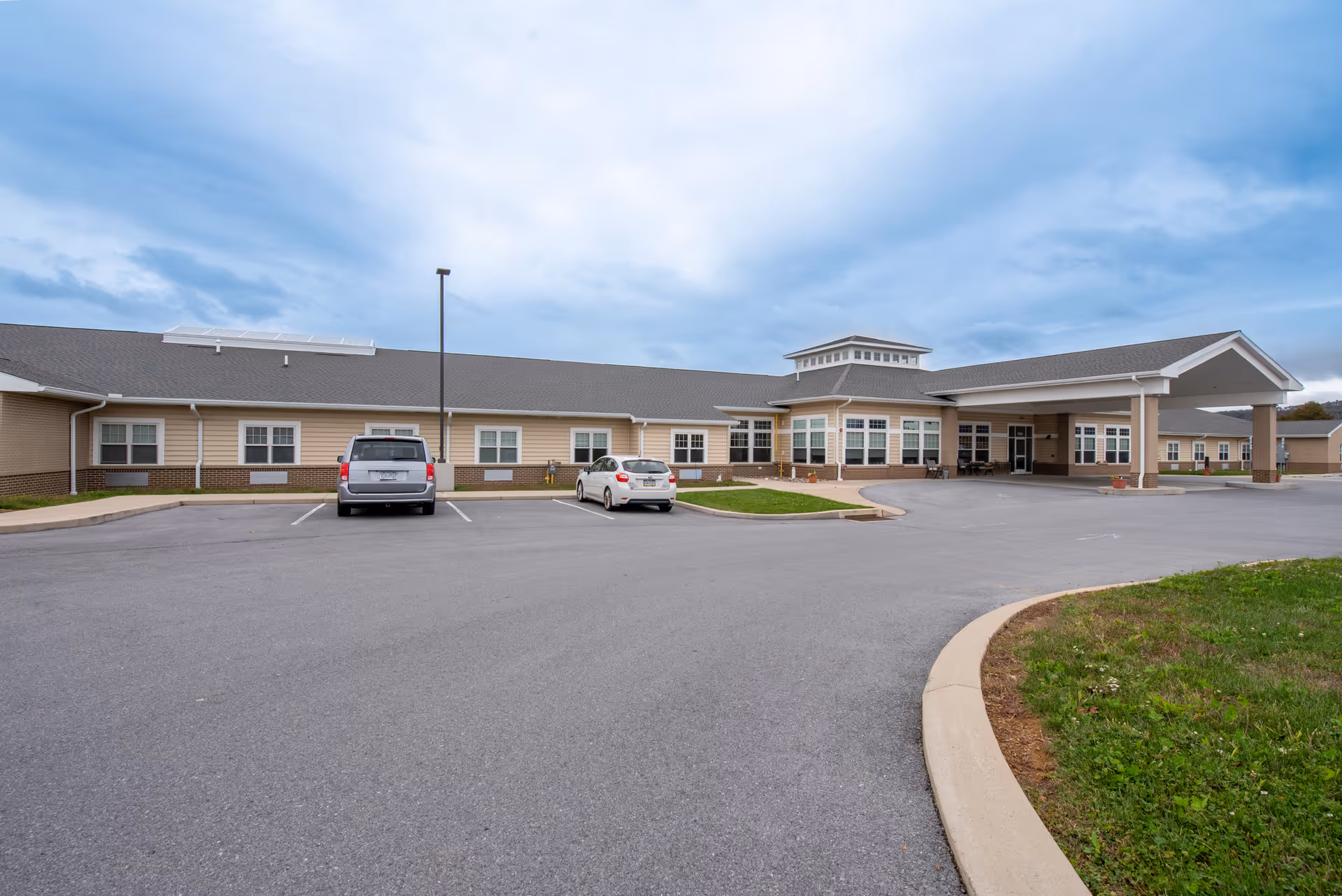 Exterior view of Wynwood House Personal Care Home showing a single-story building with beige siding and a covered entrance. There are two cars parked in the parking lot in front of the building, and the sky is partly cloudy.