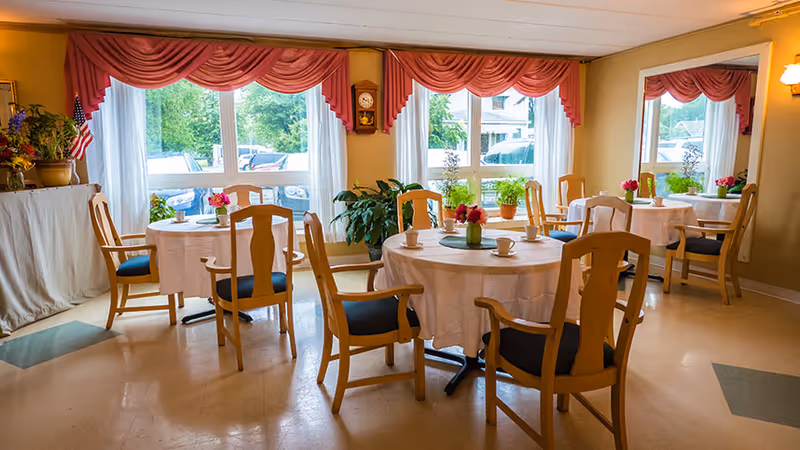 A bright dining room with round tables covered in white tablecloths, each set with cups and small flower arrangements. Wooden chairs with blue cushions surround the tables. Large windows with sheer white curtains and red valances let in natural light. There are plants on the windowsills and a wall clock between the windows. A large mirror reflects part of the room.