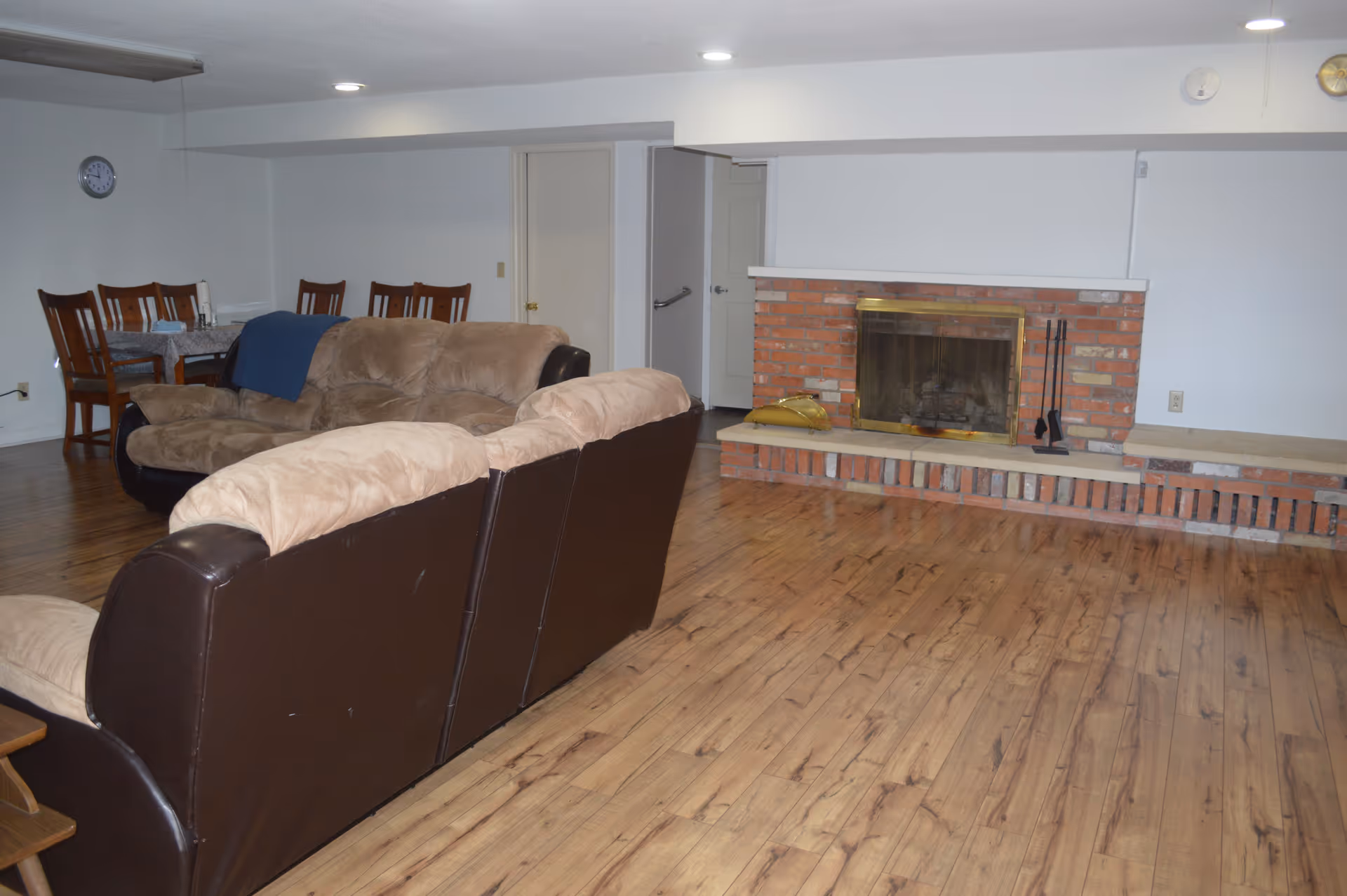A living room area with two brown and beige sofas facing a brick fireplace with a brass screen. There is a wooden dining table with six chairs in the background, a clock on the wall, and wooden flooring throughout the room.