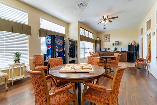 A bright common area with wooden flooring featuring a round table surrounded by six upholstered chairs with a board game on top. In the background, there are vending machines, additional seating, a kitchenette with cabinets, a refrigerator, and a ceiling fan with lights. Large windows with blinds allow natural light to fill the room.