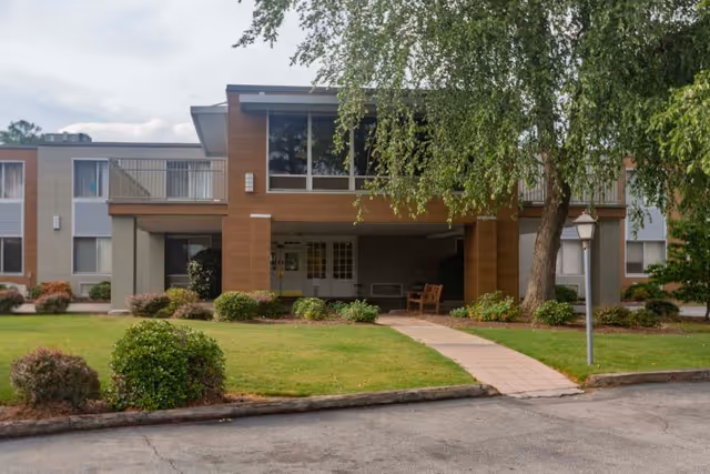 Exterior view of Millennium Post Acute Rehabilitation building with a covered entrance, large windows, a walkway leading to the entrance, green lawn, bushes, and a tree on the right side.