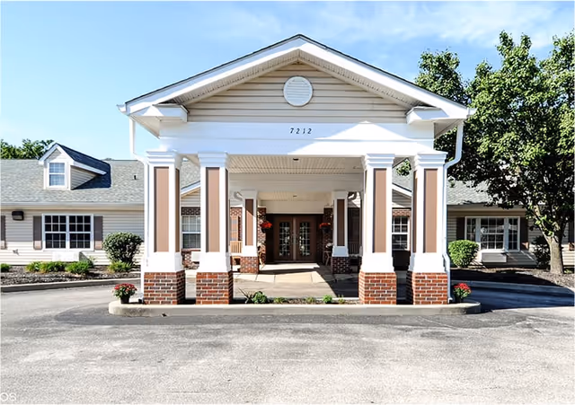 Front entrance of a senior living facility with a covered driveway supported by white columns with brick bases. The building has beige siding, multiple windows, and a tree on the right side. The address number 7212 is displayed above the entrance.