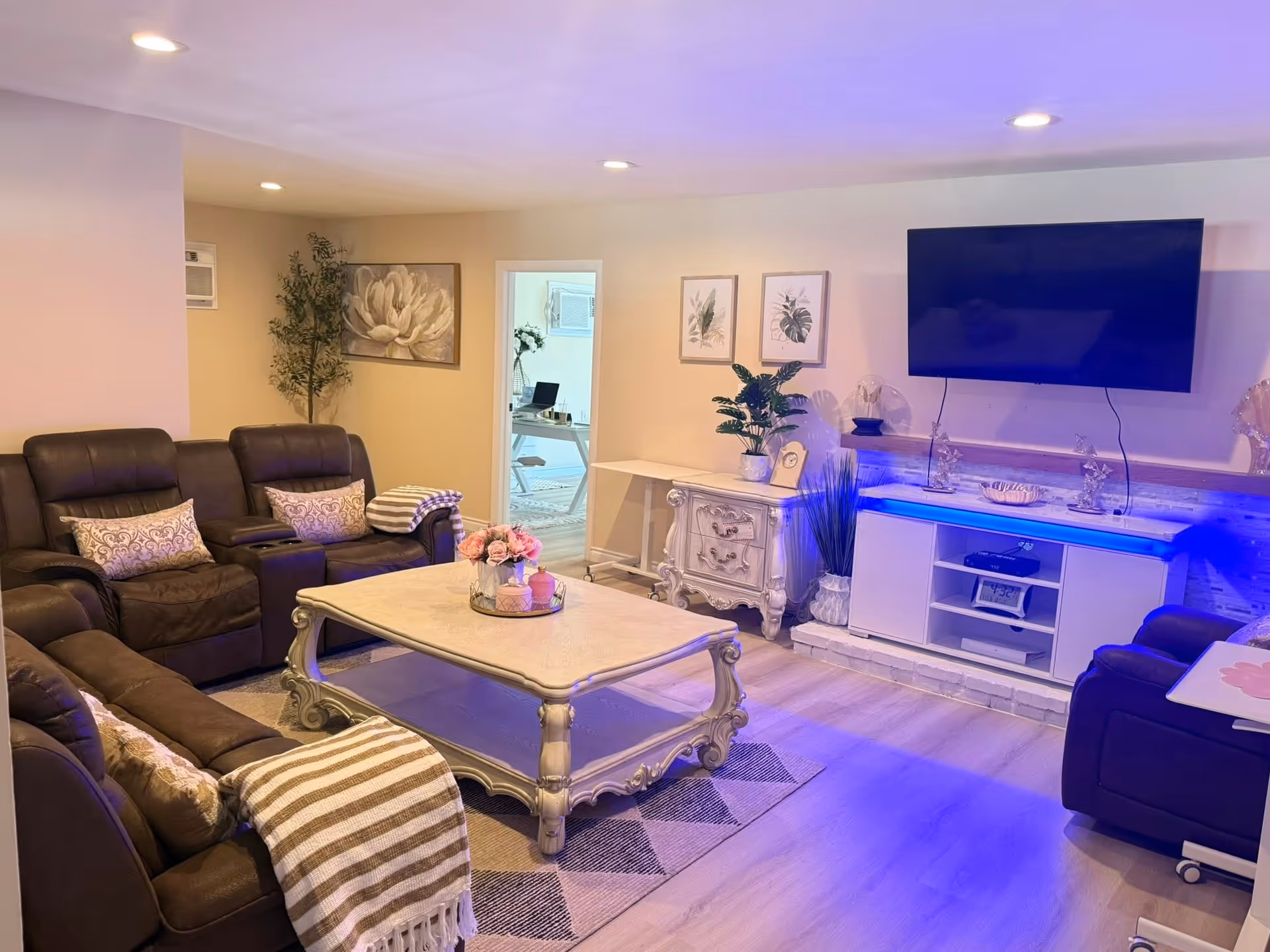 A cozy living room with brown leather recliner sofas arranged around a white ornate coffee table with a flower arrangement on top. The room features a large flat-screen TV mounted on the wall above a white entertainment unit with blue accent lighting. There are decorative plants and framed botanical prints on the walls, with a doorway leading to another room visible in the background.