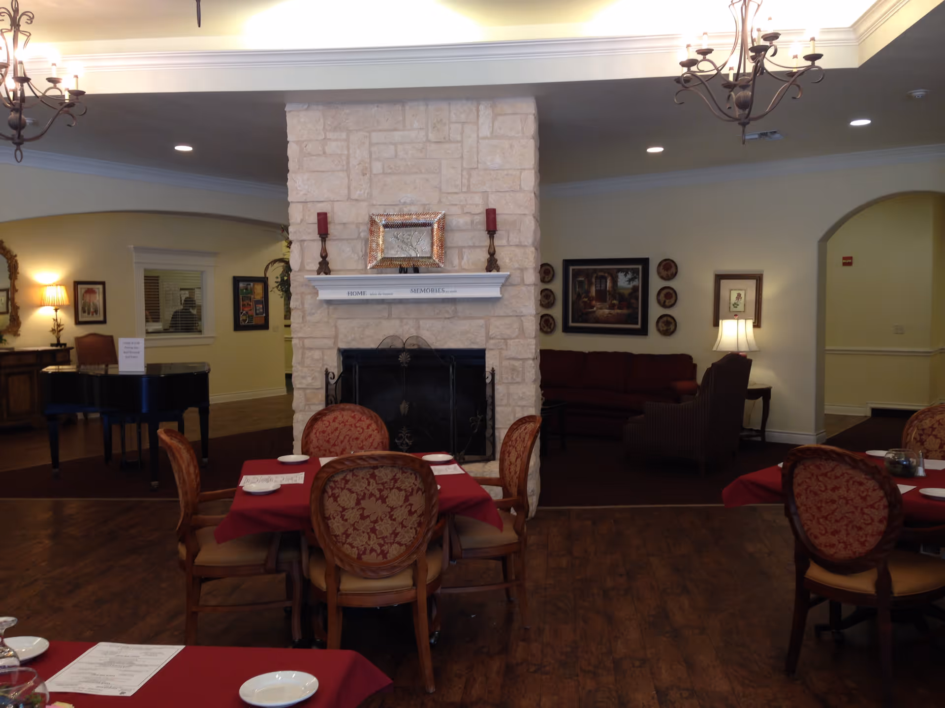 Dining room with tables covered in red tablecloths, a central stone fireplace, sofas and chandeliers.