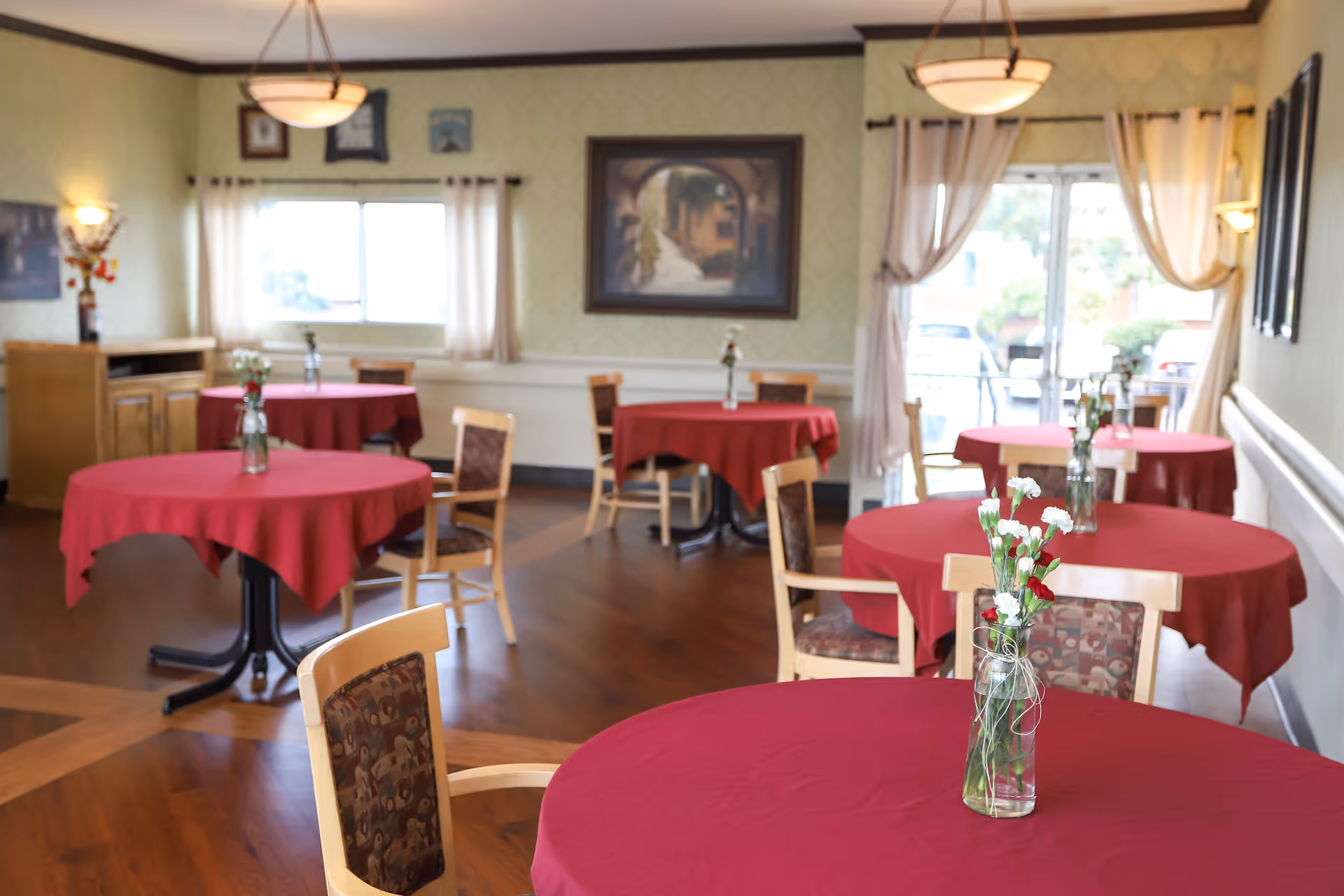A dining room with several round tables covered in red tablecloths, each with a small vase of flowers as a centerpiece. The room has wooden floors, light-colored walls with framed artwork, and windows with curtains letting in natural light.