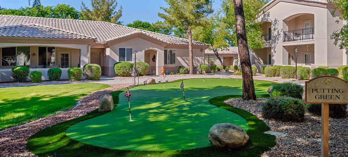 Outdoor putting green area with several small flags on the green, surrounded by rocks and bushes, with senior living facility buildings in the background under a clear blue sky.