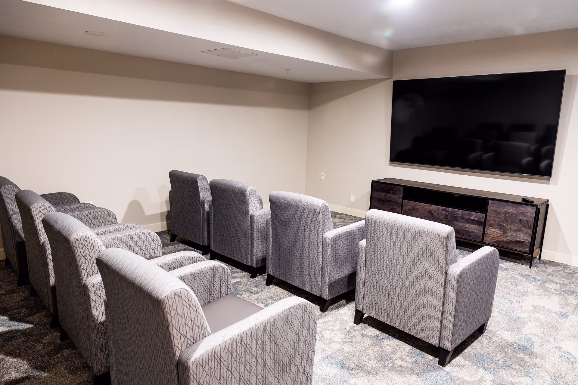 A small theater room with two rows of gray patterned armchairs facing a large flat-screen TV mounted on a beige wall. Below the TV is a dark wooden media console. The room has a carpeted floor with a subtle pattern and a low ceiling.