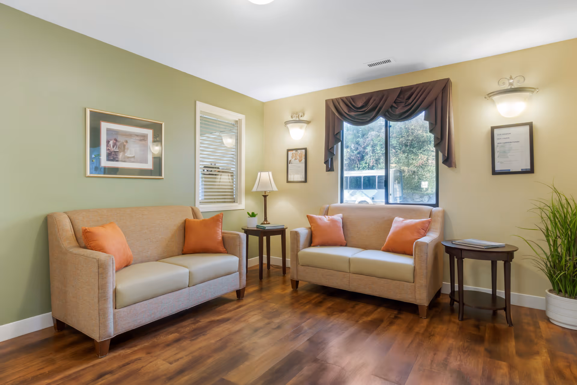 A cozy living room area with two beige sofas, each adorned with two orange throw pillows. Between the sofas are two small wooden side tables, one with a lamp and a small plant, and the other with a closed book. The room has wooden flooring, light green and beige walls, a window with brown drapes, framed pictures on the walls, and a potted plant in the corner.