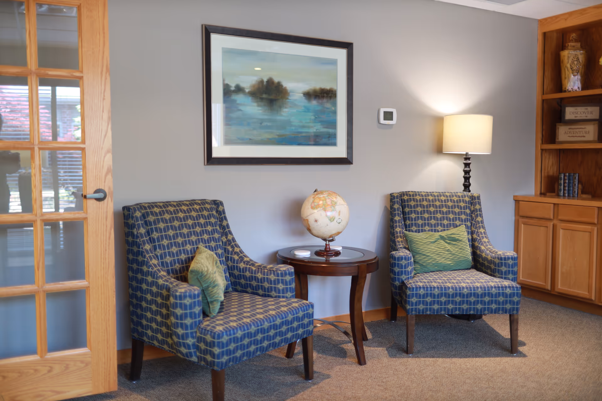 A cozy sitting area with two patterned blue armchairs each with a green cushion, a round wooden side table between them holding a globe, a table lamp on the right side, a framed painting of a lake scene on the wall, and a wooden door with glass panes on the left.