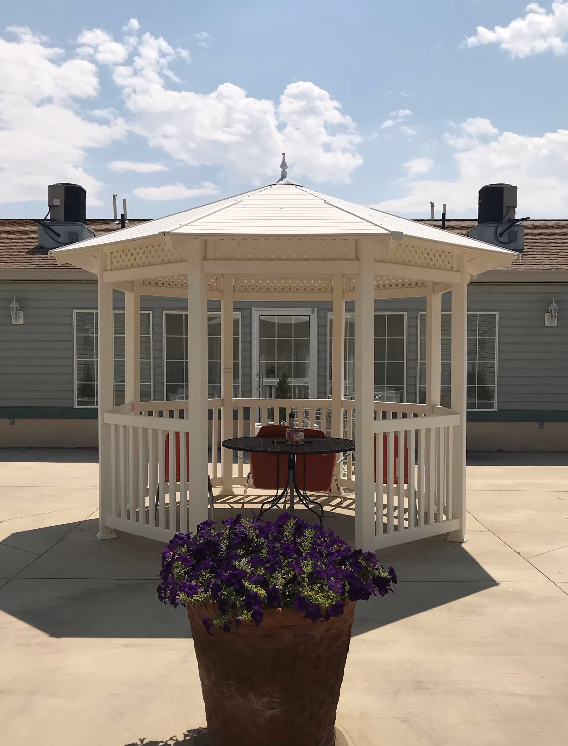 Outdoor patio area at GoodLife Senior Living & Memory Care of Elmo, UT featuring a white gazebo with a round black table and red chairs inside. In the foreground, there is a large basket planter filled with purple flowers. The building with multiple windows is visible in the background under a partly cloudy sky.