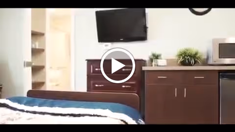 Interior view of a senior living facility bedroom featuring a bed with a blue and white blanket, a flat-screen TV mounted on the wall, a wooden dresser, a countertop with a microwave, and a small plant. A doorway leads to a bathroom area.