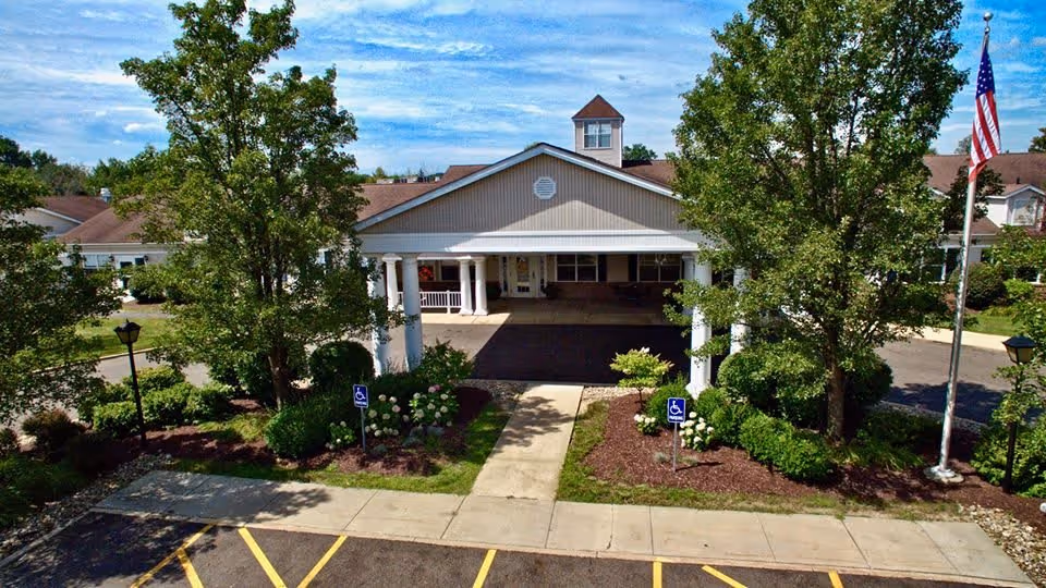 Front exterior view of a single-story building with a covered entrance supported by white columns, surrounded by trees and landscaping. There are two handicap parking signs near the entrance and an American flag on a flagpole to the right.