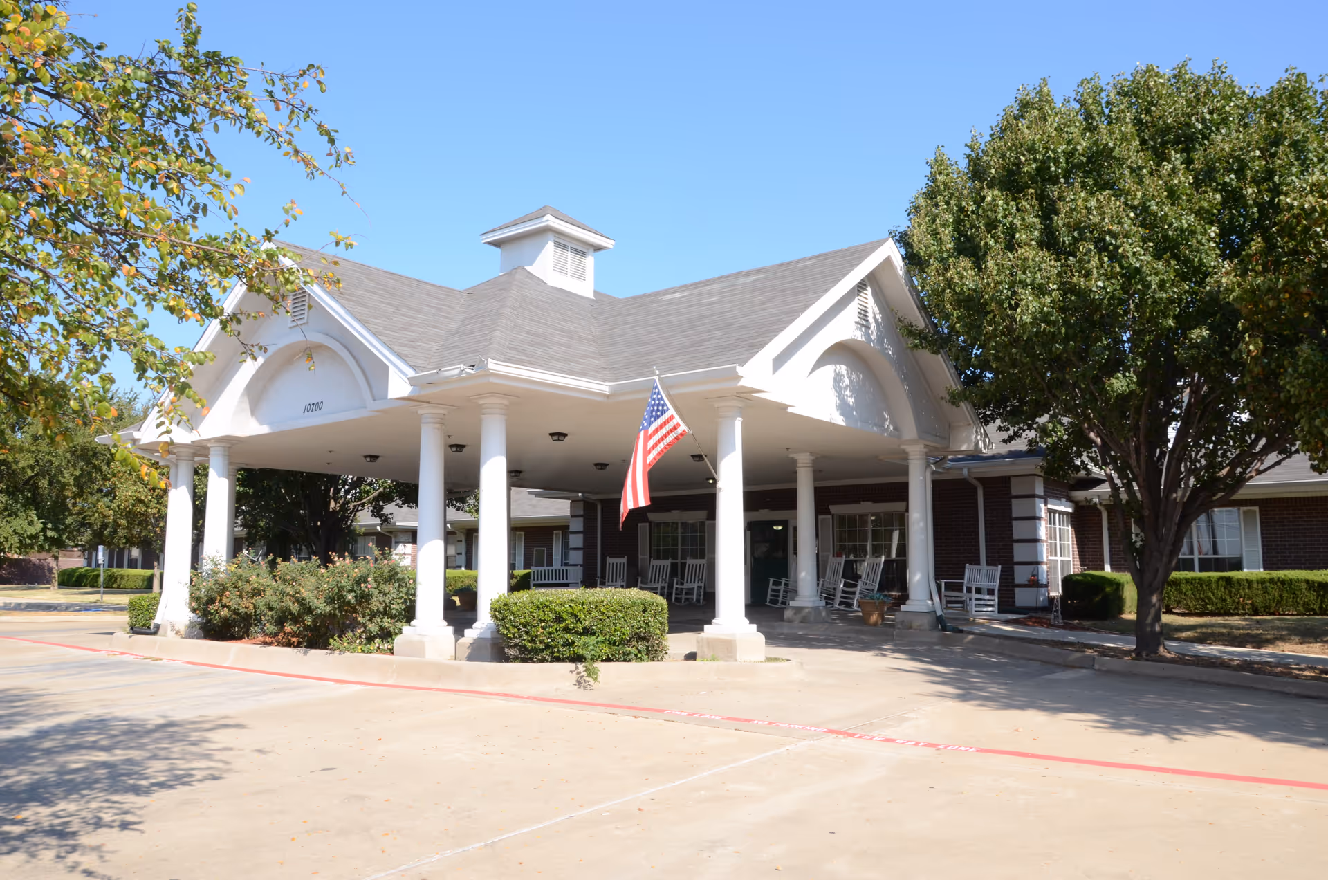 Front exterior view of a senior living facility building with a covered entrance supported by white columns, an American flag hanging from the roof, rocking chairs on the porch, and trees and bushes surrounding the area under a clear blue sky.