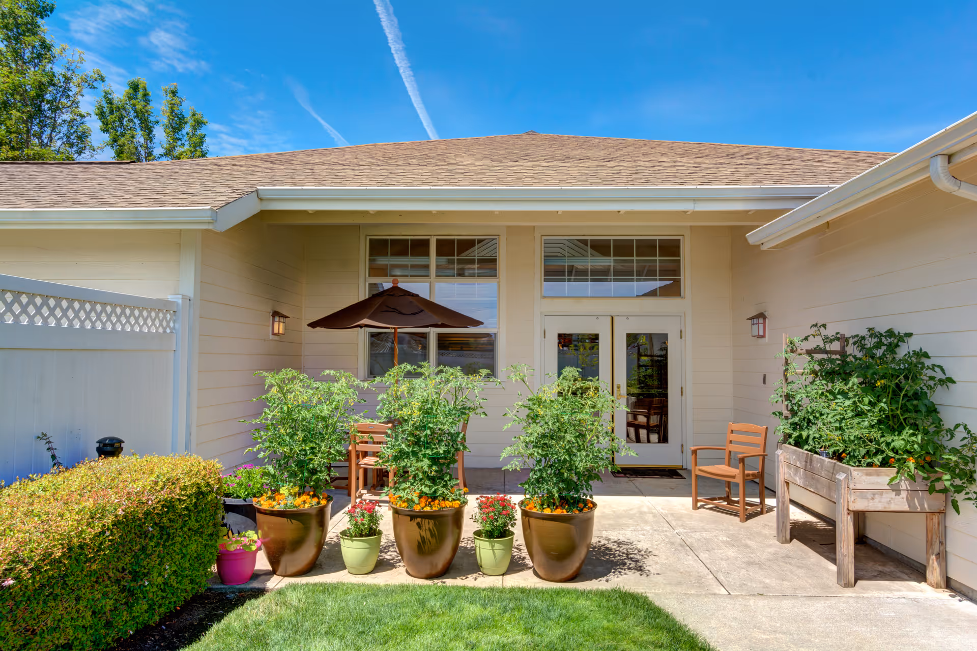 Outdoor patio area at Rosewood Memory Care with potted plants, a wooden chair, a table with an umbrella, and a raised garden bed against the building exterior under a clear blue sky.