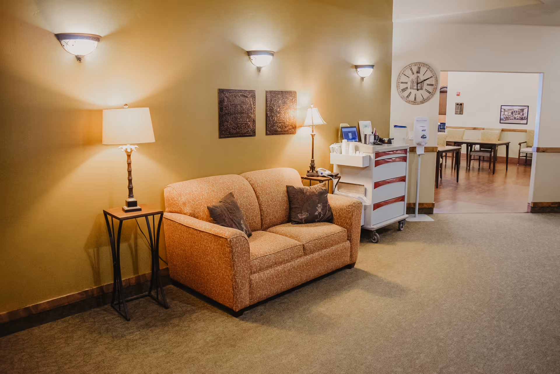 A cozy seating area in an assisted living facility with a patterned orange loveseat, two decorative pillows, a side table with a lamp, and wall-mounted lights. In the background, there is a hallway leading to a dining area with tables and chairs, a large wall clock, and a medical cart with supplies.