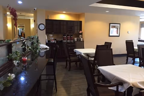 Interior view of a dining area in a senior living facility with tables covered in white tablecloths, dark wooden chairs, a counter with decorative plants and candles, and a cabinet with coffee dispensers against a beige wall.