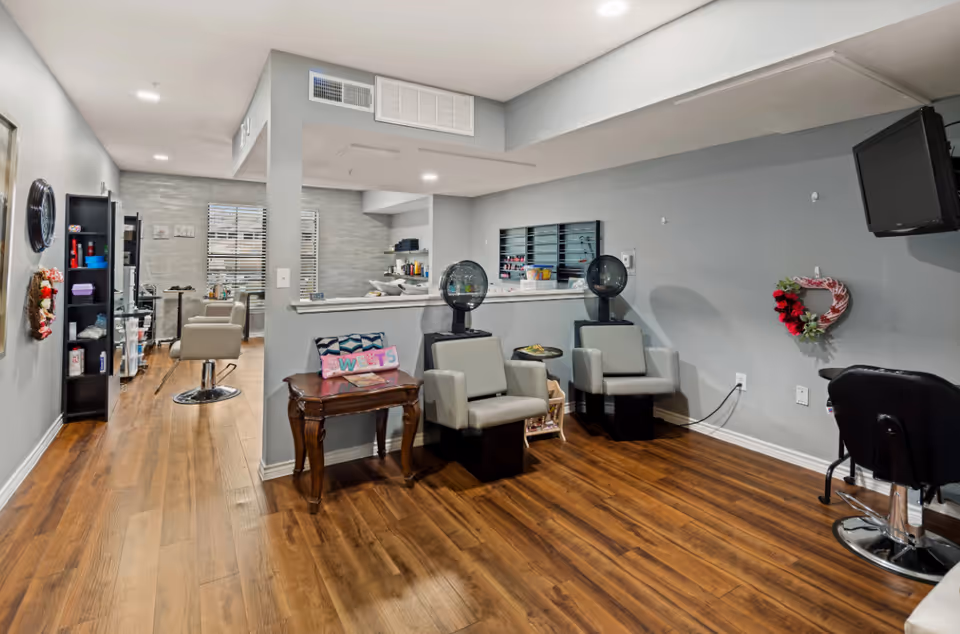 Interior view of a hair salon area in a senior living facility with wooden flooring, two hair drying chairs with hooded dryers, a small wooden table with a decorative pillow, salon chairs, shelves with hair products, and a wall-mounted TV.