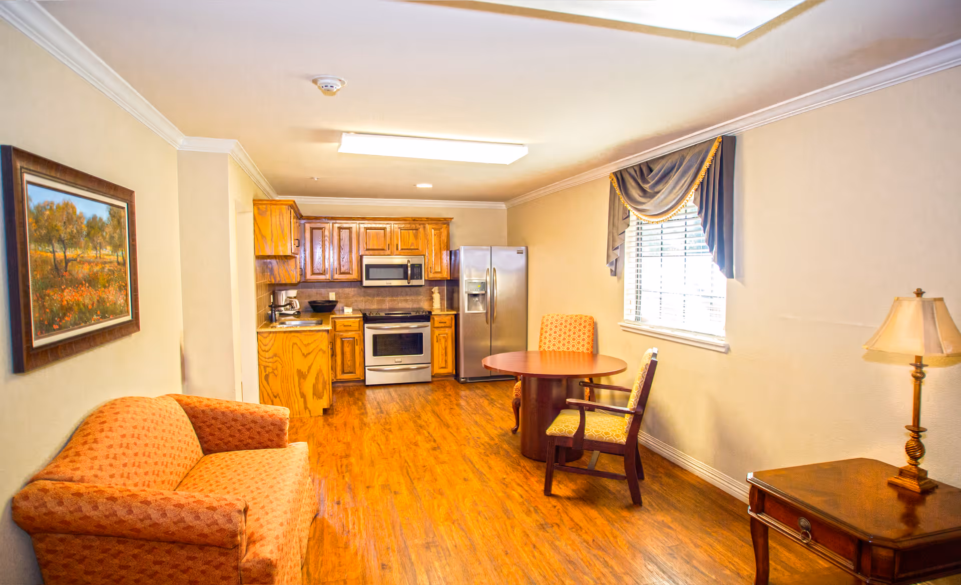Interior view of a room in Sandy Lake Rehabilitation and Care Center featuring a small kitchen area with wooden cabinets, a stainless steel refrigerator, microwave, and stove. The room also includes a round wooden table with two upholstered chairs, an orange patterned sofa, a wooden side table with a lamp, and a window with draped curtains. A framed landscape painting hangs on the wall above the sofa.