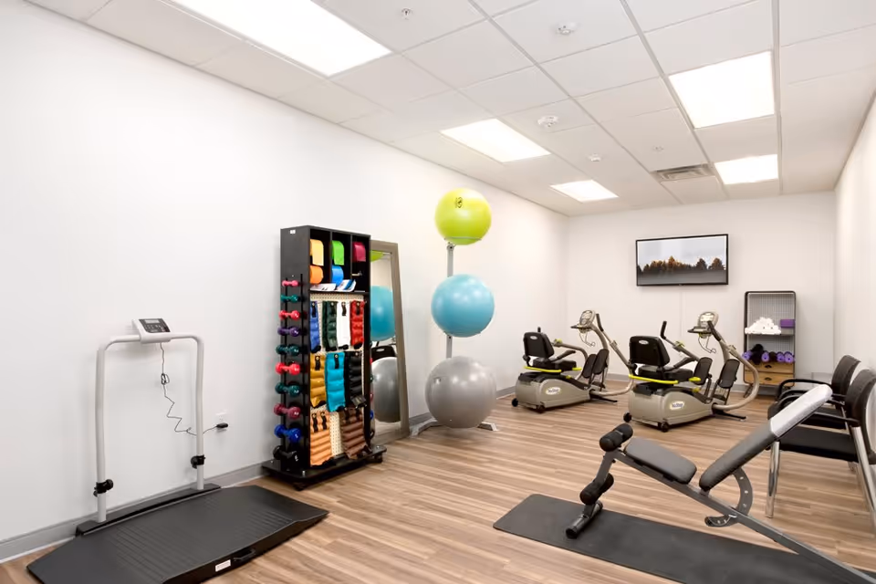A small fitness room with exercise equipment including a treadmill, two recumbent bikes, a weight bench, a rack with colorful dumbbells and resistance bands, three large exercise balls stacked vertically, a full-length mirror, and a shelf with rolled towels and yoga mats. The room has white walls, a wood floor, and a ceiling with fluorescent lights. A framed picture of trees is mounted on the far wall.