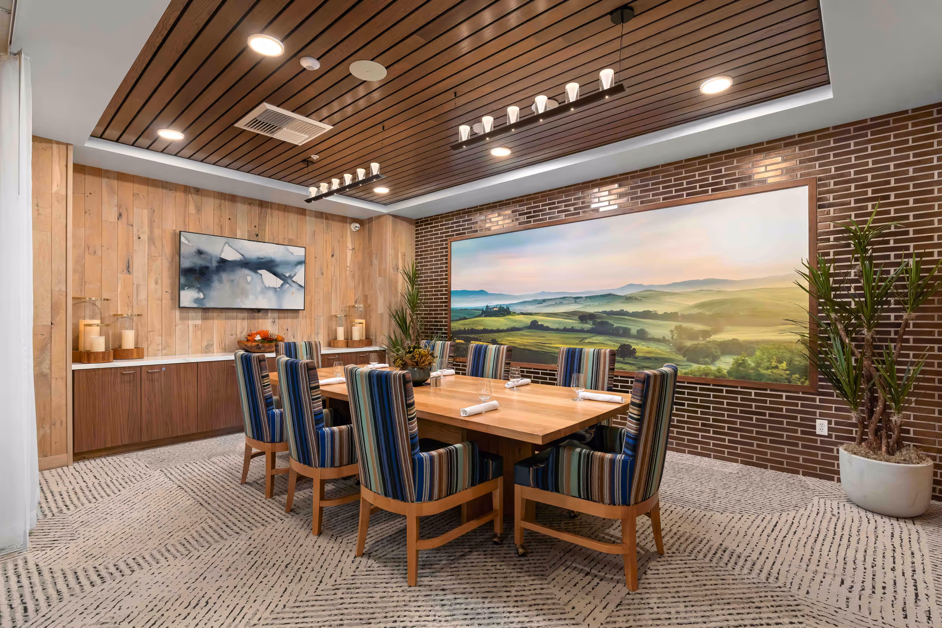 A dining room with a large wooden table surrounded by eight striped upholstered chairs. The room features a wood-paneled ceiling with modern light fixtures, a brick accent wall with a large landscape painting, a wooden sideboard with candles and decorative items, and a potted plant in the corner.