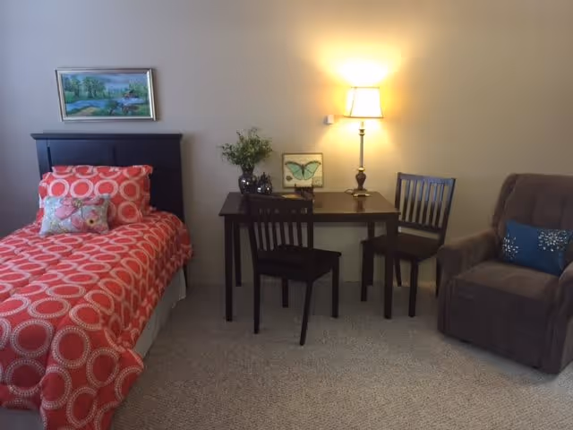 A cozy bedroom featuring a single bed with a red and white circular patterned bedspread and a decorative pillow. Next to the bed is a dark wooden table with two chairs, a table lamp, a small framed butterfly picture, and a vase with flowers. To the right is a comfortable brown armchair with a blue pillow. A framed landscape painting hangs on the wall above the bed.