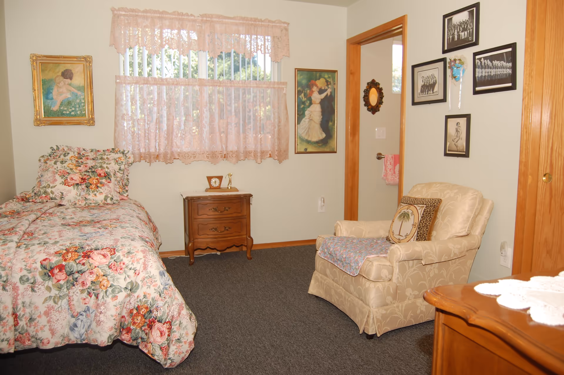 A cozy bedroom with a floral bedspread on a single bed, a wooden nightstand with a clock and small figurine, a beige upholstered armchair with decorative pillows and a small blanket, and several framed pictures and a small mirror on the walls. The window has pink lace curtains.
