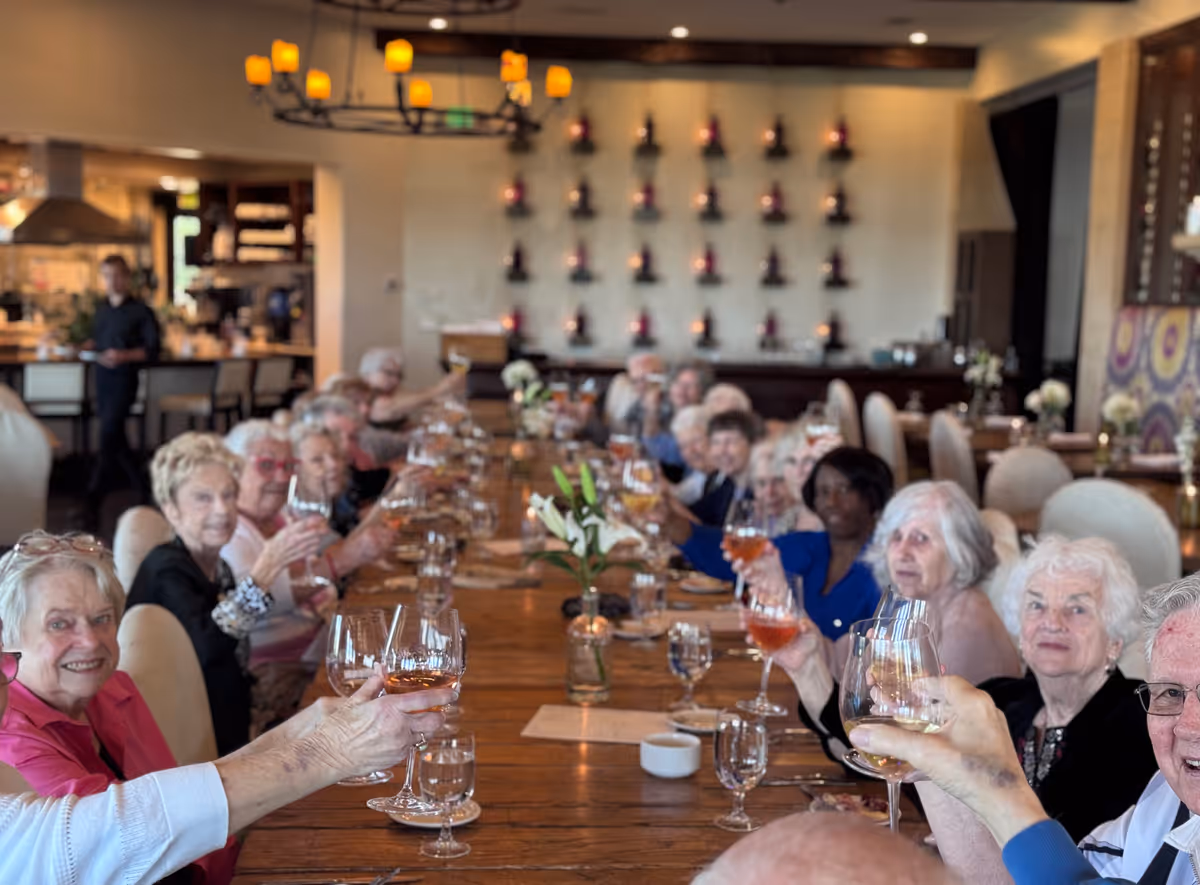 A group of elderly people sitting around a long wooden dining table in a well-lit restaurant or dining room, raising their glasses in a toast. The background shows a wall with decorative lighting and a waiter standing near the kitchen area.