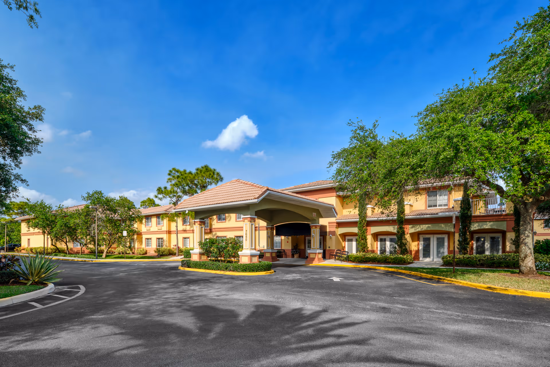 Front entrance of a two-story senior living building with a covered porte-cochere, driveway, and surrounding trees under a blue sky.