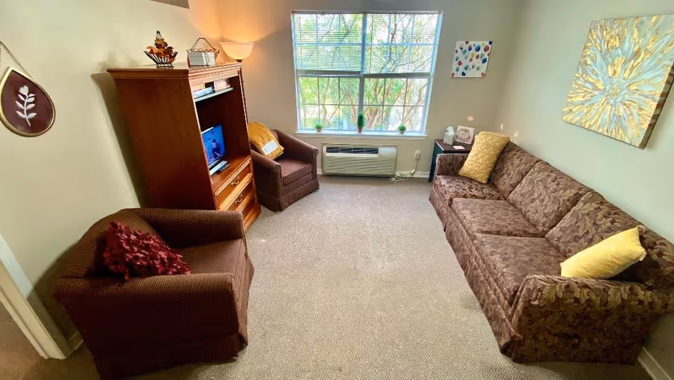 A cozy living room in a senior living facility with a patterned brown sofa and two brown armchairs, one with a red pillow and the other with a yellow pillow. There is a wooden entertainment center with a small TV, a window with a view of greenery outside, and wall decorations including a leaf-shaped plaque and two pieces of artwork. The room is carpeted and softly lit by a floor lamp.