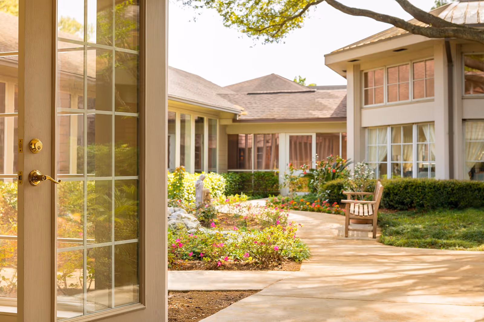 View through a glass-paned door looking out onto a garden courtyard with a paved walkway, colorful flower beds, green shrubs, a wooden bench, and a building with large windows and beige walls under a sunny sky.