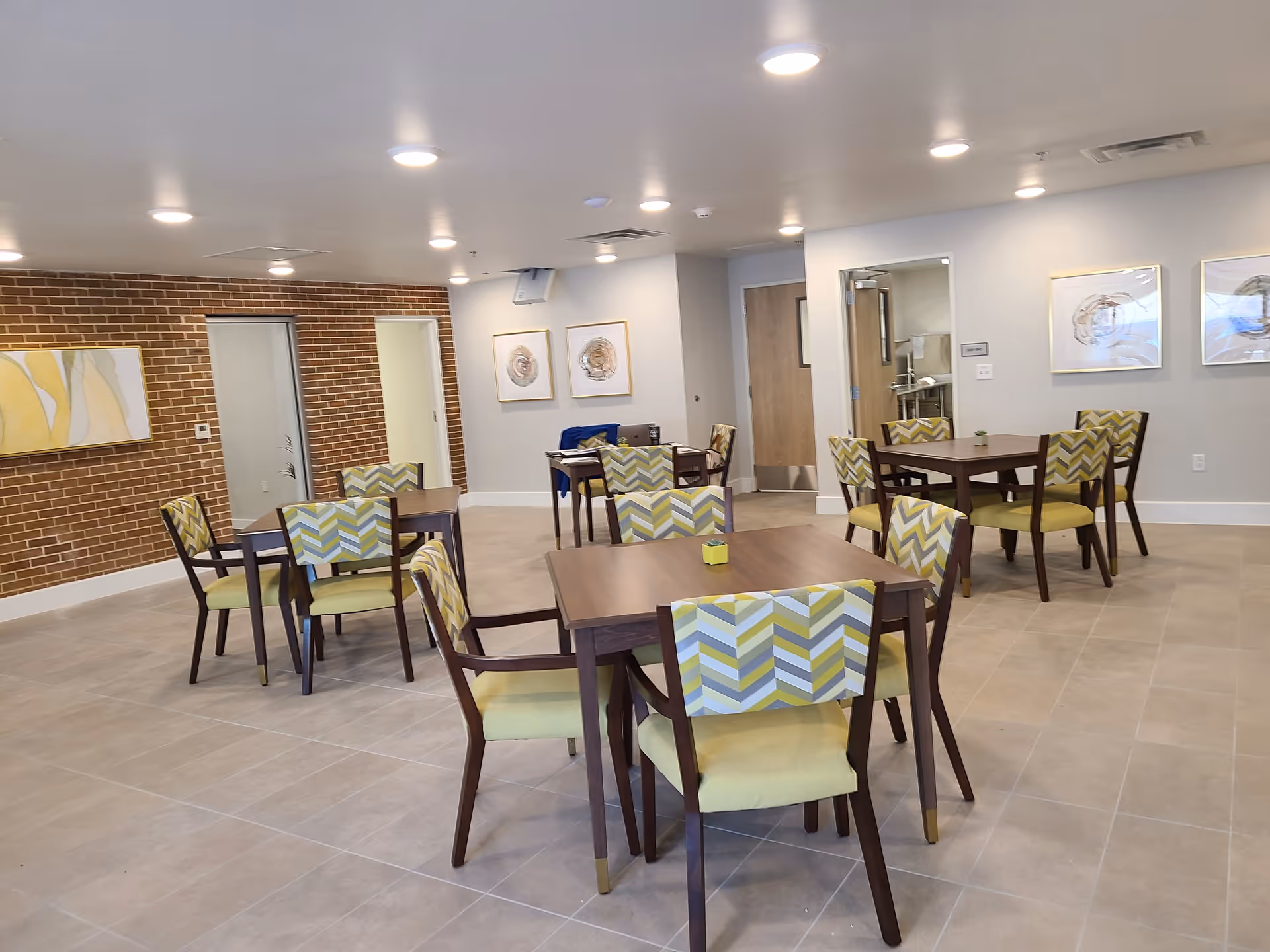 A bright dining room with several wooden square tables and chairs featuring green and patterned upholstery. The room has a tiled floor, white walls, and a brick accent wall with framed artwork. Ceiling lights illuminate the space, and there is a door leading to another room in the background.