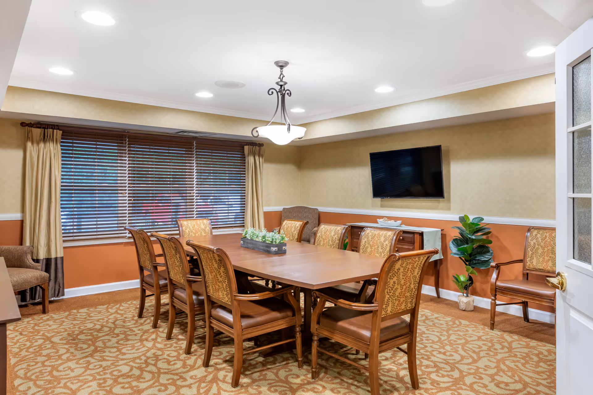 A well-lit dining room with a long wooden table surrounded by eight upholstered chairs. The room features a patterned carpet, beige and orange walls with white trim, a large window with closed blinds and curtains, a wall-mounted flat-screen TV, a small cabinet, a potted plant, and a ceiling light fixture.