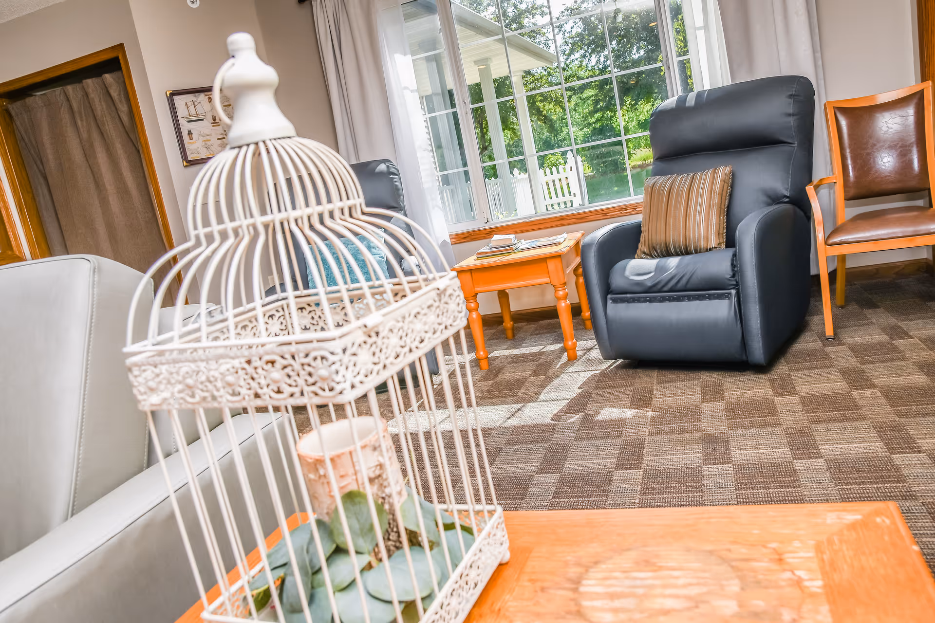 A cozy living room with a decorative white birdcage on a coffee table, a black recliner and wooden chairs by a large window.