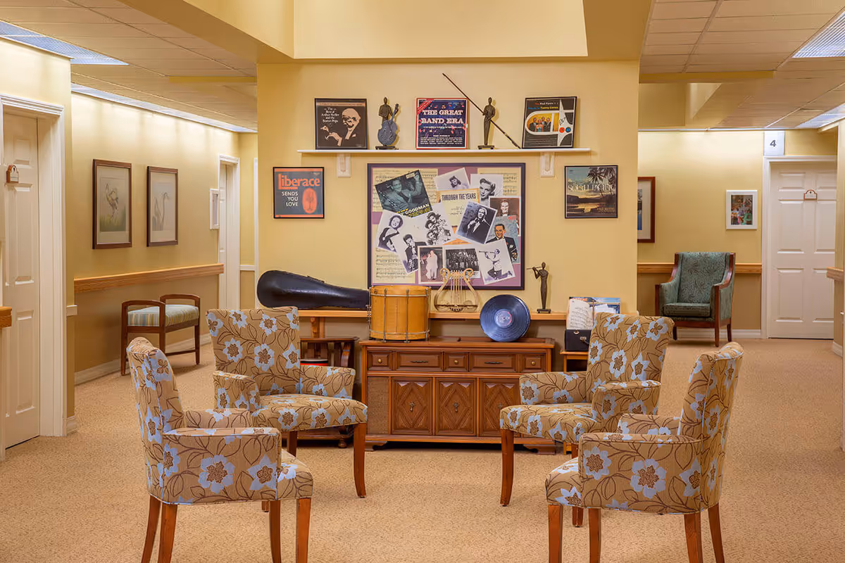 A cozy common area in a senior living facility with four floral-patterned armchairs arranged in a circle on a beige carpet. Behind the chairs is a wooden cabinet topped with musical decorations including a drum, a lyre, a vinyl record, and small statues. The wall above the cabinet features framed pictures and posters related to music. The hallway has cream-colored walls with framed artwork and several white doors.