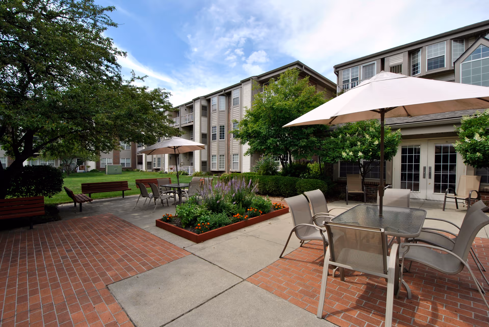 Outdoor patio area at Westhaven Manor featuring tables with umbrellas, chairs, benches, a flower bed, and a multi-story residential building in the background under a partly cloudy sky.