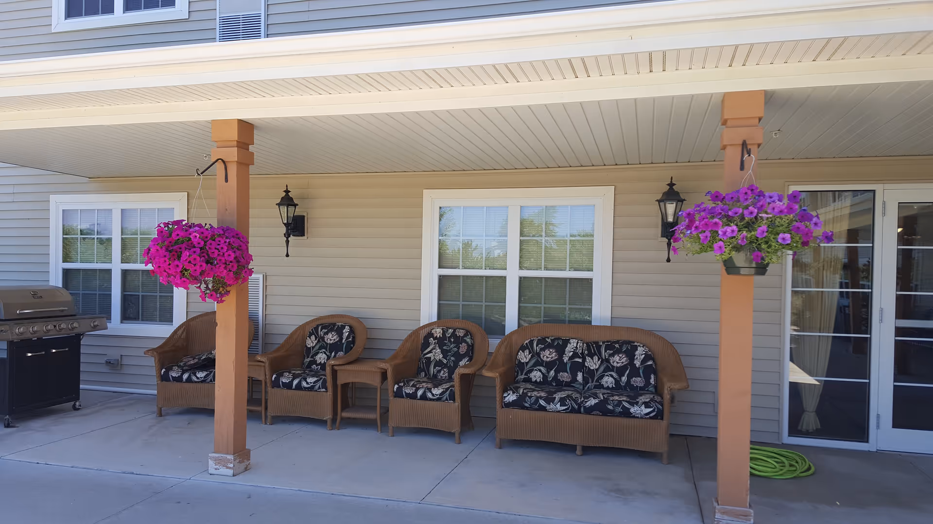 Outdoor covered patio area with wicker seating including a loveseat, two chairs, and a small table. Two hanging baskets with vibrant purple flowers are suspended from wooden posts. A grill is positioned on the left side near a window, and a green garden hose is coiled on the ground near a glass door on the right.