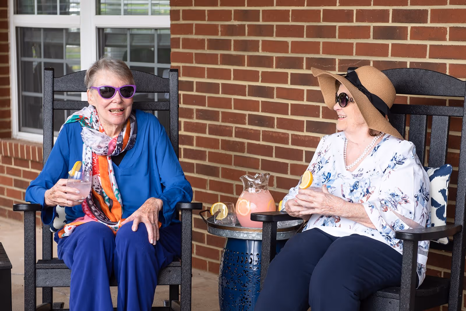 Two elderly women sitting on black rocking chairs on a patio against a brick wall. Both are wearing sunglasses; one woman is dressed in a blue top and colorful scarf, holding a glass of pink lemonade with a lemon slice, while the other woman wears a floral blouse and wide-brimmed hat, also holding a glass of pink lemonade. A small round table between them holds a pitcher of pink lemonade with lemon slices.