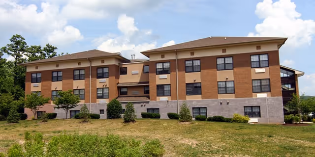 Exterior view of a three-story senior living facility building with a combination of brick and stone facade, multiple windows, and a sloped roof, surrounded by grass and small trees under a partly cloudy sky.