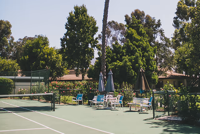 Outdoor tennis court with a seating area including chairs, tables, and umbrellas surrounded by trees and greenery under a clear sky.