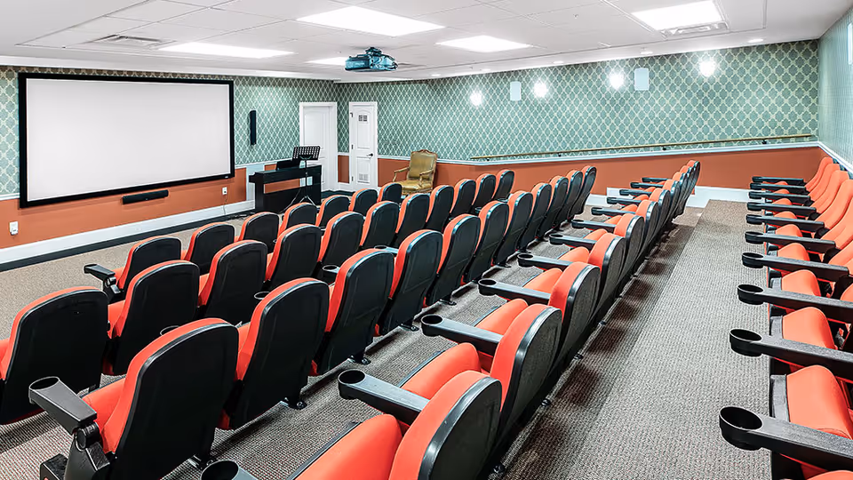 A small theater room with rows of red and black cushioned seats facing a large white projection screen on the wall. The room has patterned green wallpaper with a reddish-brown lower wall section, carpeted floor, ceiling lights, and a projector mounted on the ceiling. There is also a music stand and a single armchair near the back wall.